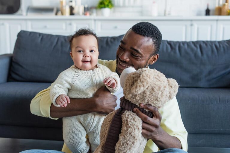 Bebê negro com roupa de lã sorri no colo de um homem negro.